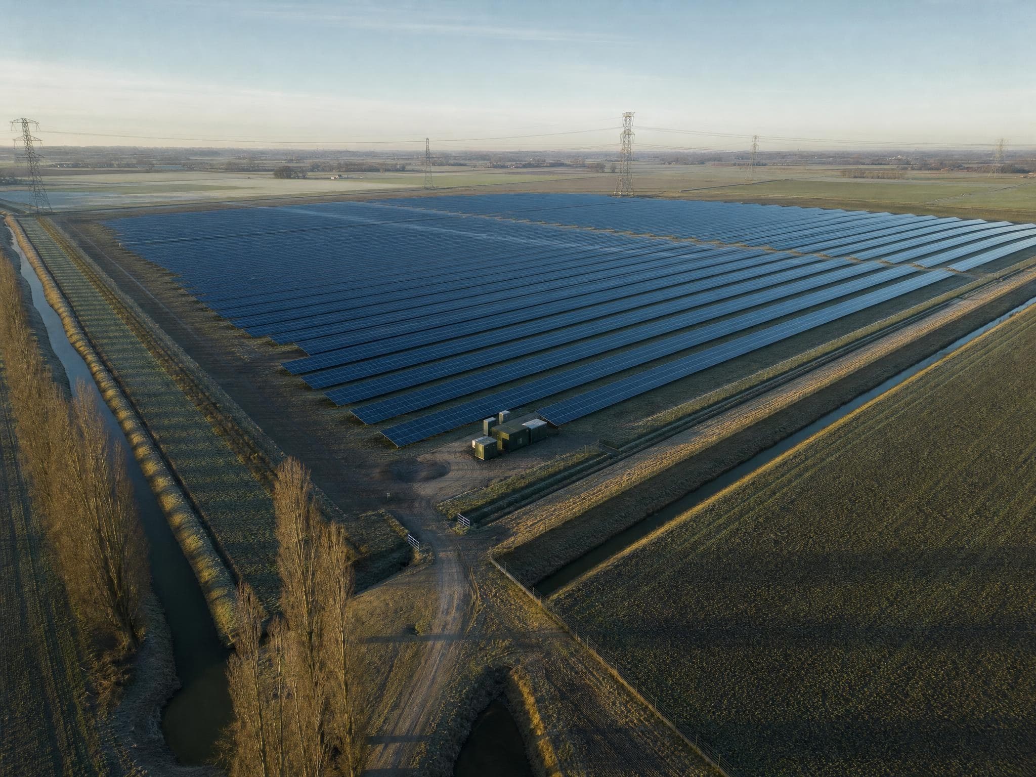 Luftbild eines Solarparks in flacher Landschaft, im Hintergrund eine Hochspannungsleitung